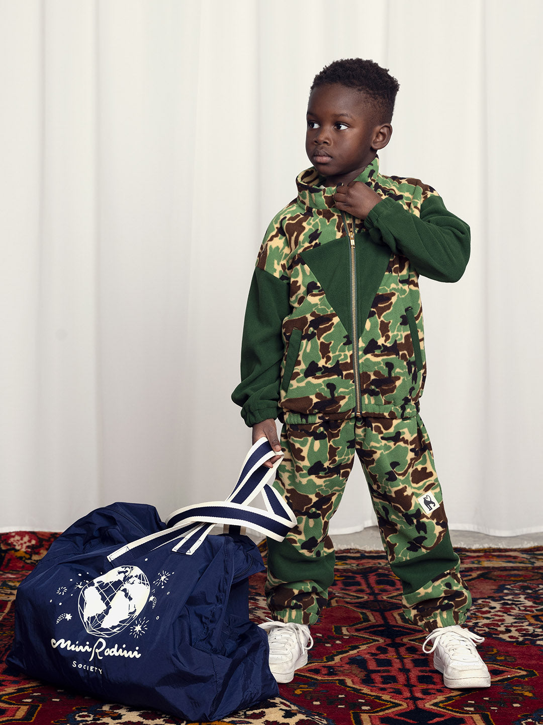 Kid wearing green fleece camouflage tracksuit carrying a large navy blue sports bag with world illustration 