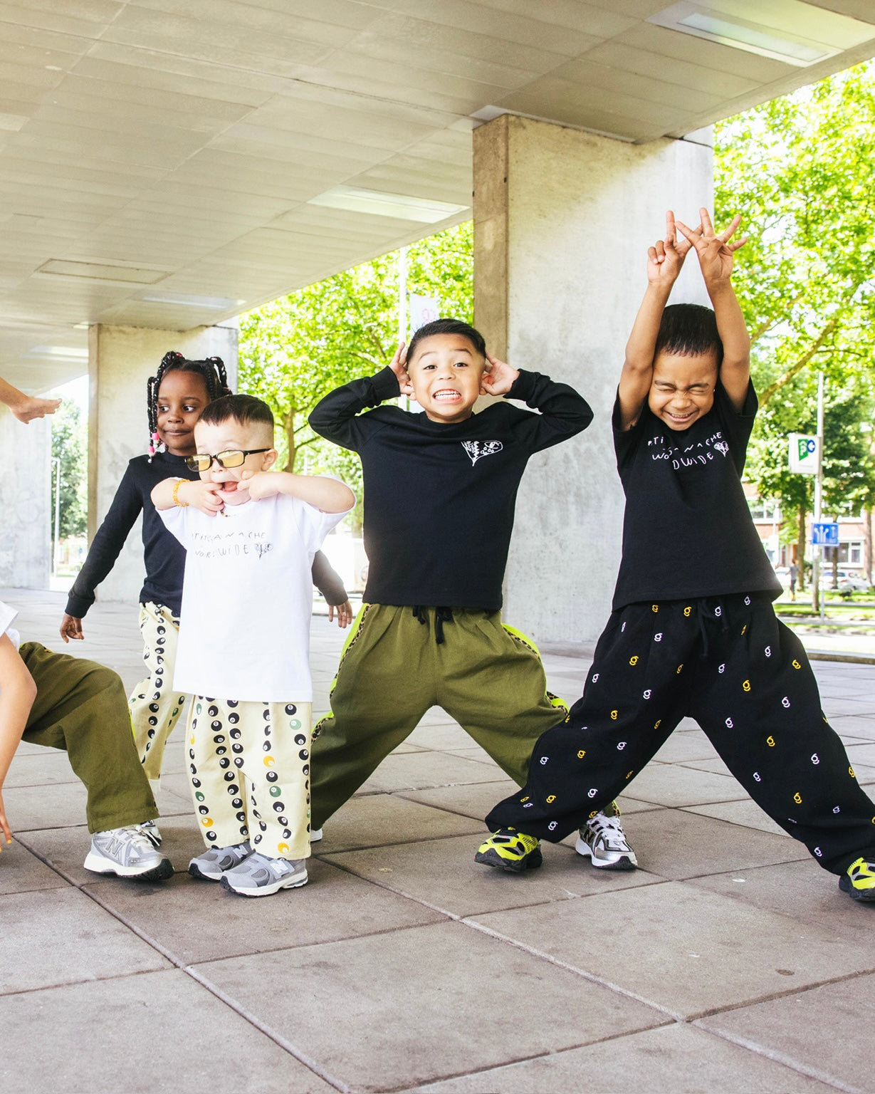 4 children making funny faces in a outdoor urban area. Weaing oversized trousers in black with small logos, khaki green with neon yellow and leopard print and yellow 8 ball printed trousers