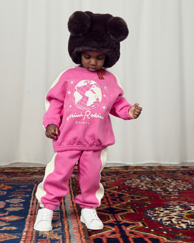 Small child weaing pink sweatshirt and sweatpnts with white stripe and world illustration. With white sneakers and a brown fur hood.